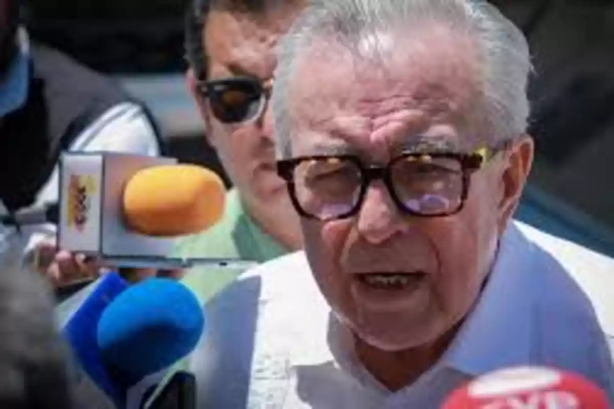 An older man with glasses speaks in front of several microphones of different colors.