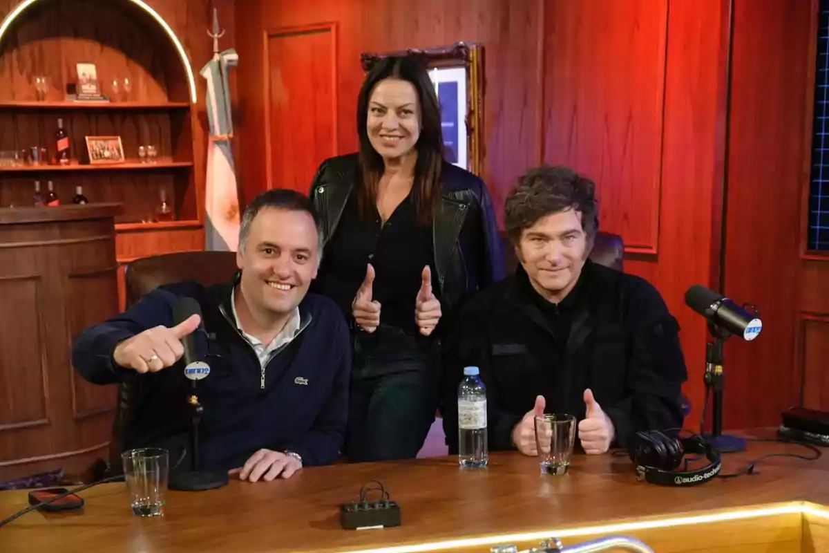 Three people pose smiling in a recording studio with microphones and an Argentine flag in the background.
