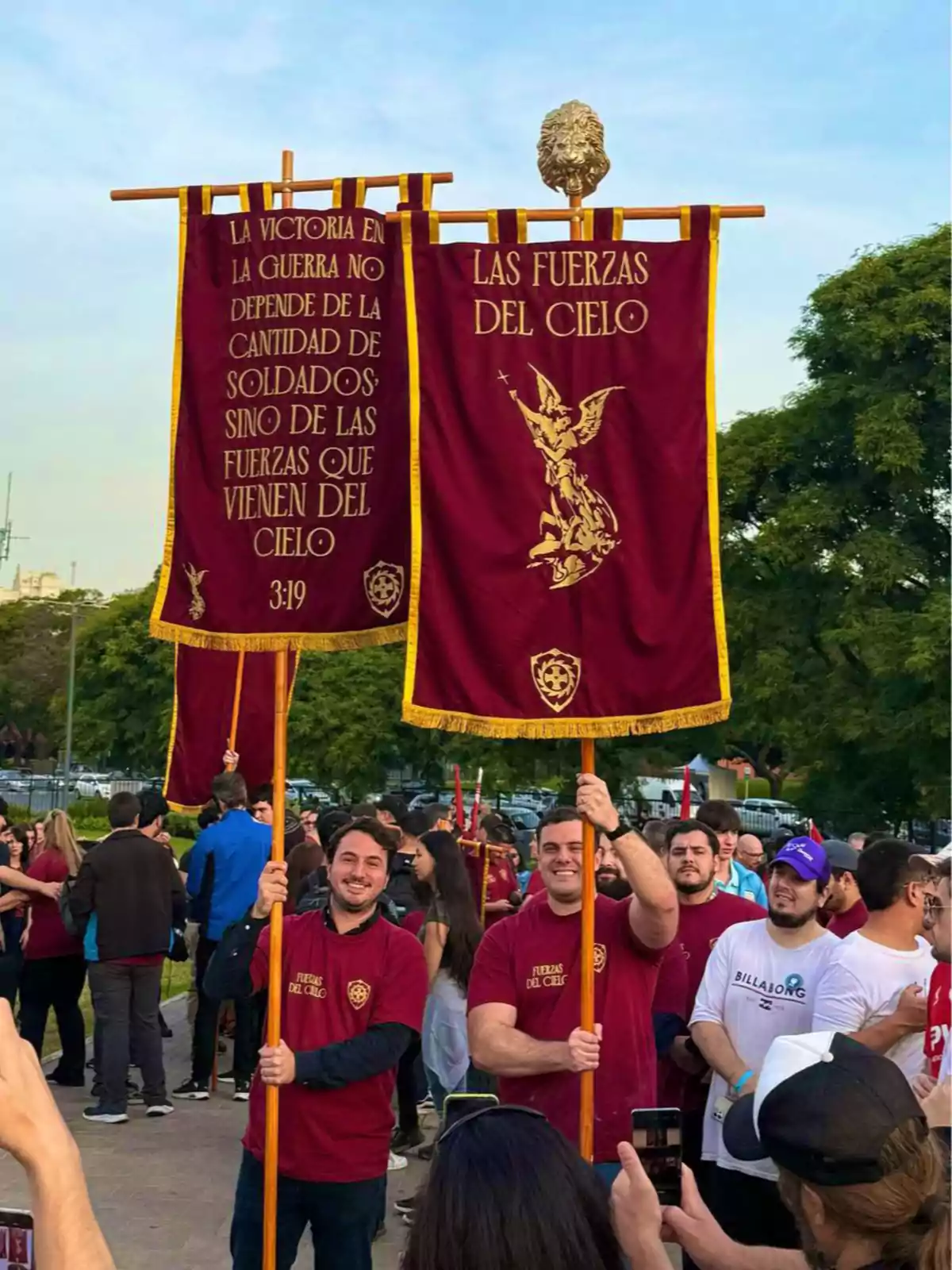 Un grupo de personas sostiene estandartes rojos con inscripciones doradas en un evento al aire libre. Un grupo de personas sostiene estandartes rojos con inscripciones doradas en un evento al aire libre.