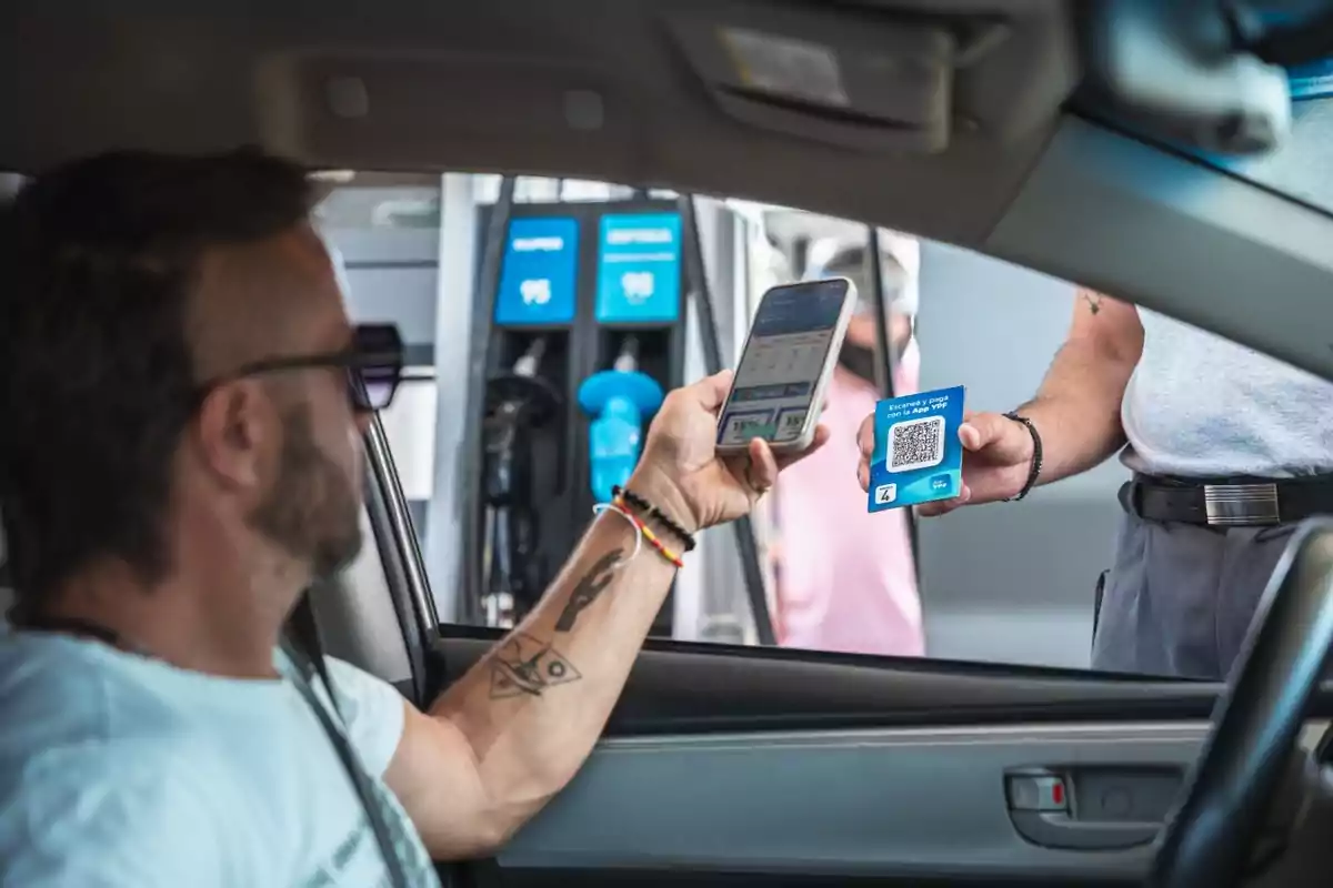 A man inside a car holds a mobile phone while another person outside shows him a card with a QR code at a gas station.