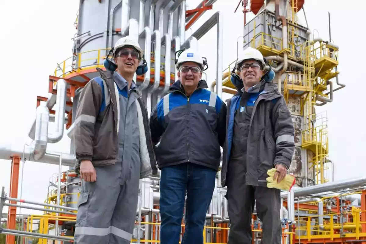 Tres trabajadores con casco y equipo de seguridad posan sonrientes frente a una estructura industrial con tuberías y plataformas amarillas