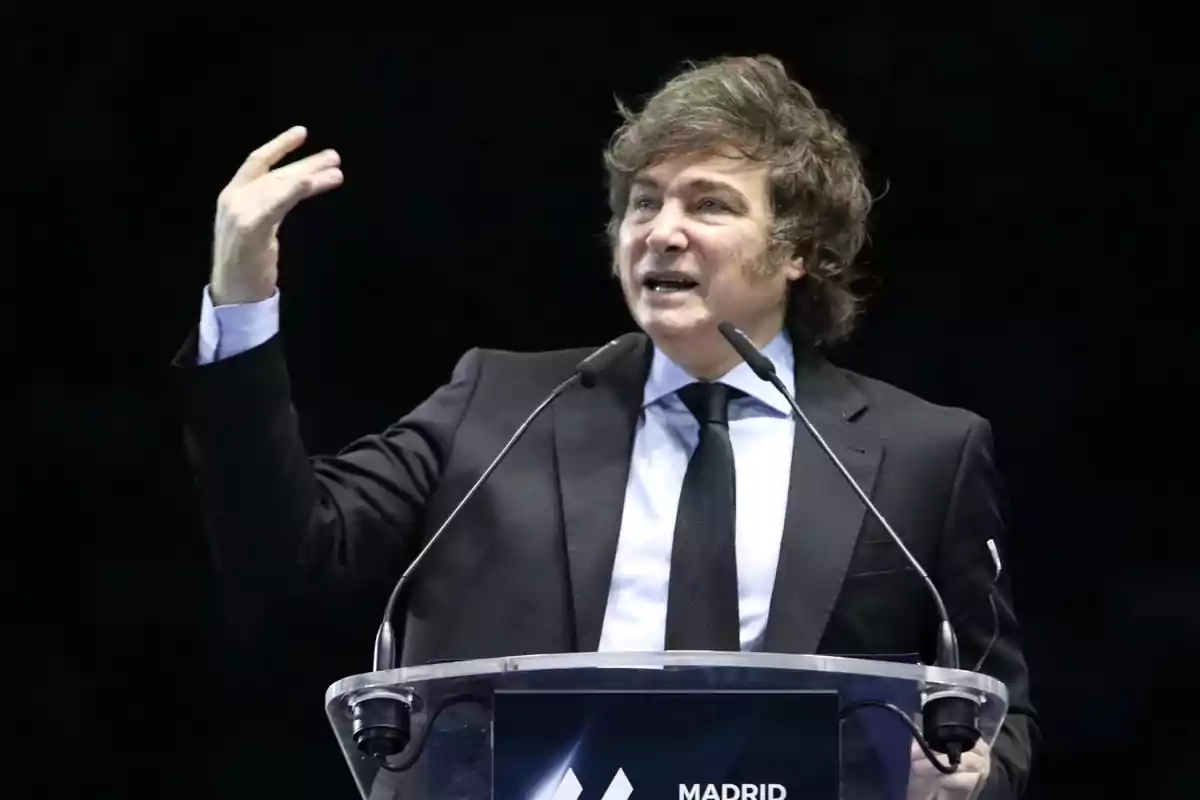 Brown-haired man in a dark suit speaking at a podium with two microphones and gesturing with his hand Brown-haired man in a dark suit speaking at a podium with two microphones and gesturing with his hand