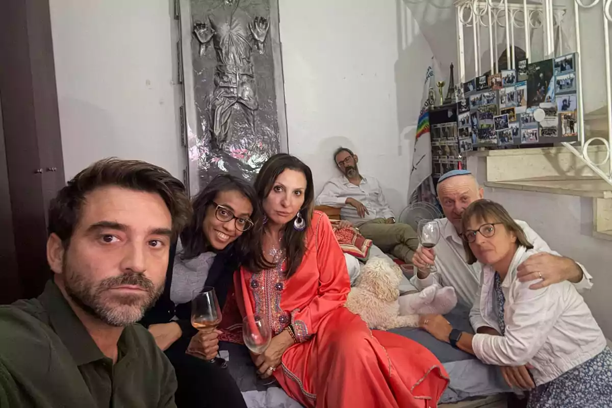 Un grupo de seis personas posa en una habitación decorada con fotos y un relieve en la pared, algunas sostienen copas de vino y hay un perro blanco acostado en la cama