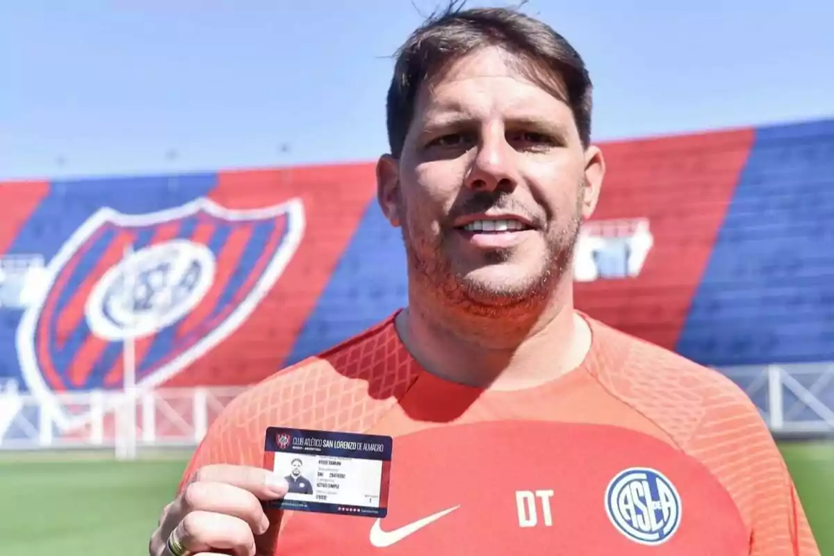 Man wearing a San Lorenzo jersey holding a credential in a stadium with red and blue stands in the background Man wearing a San Lorenzo jersey holding a credential in a stadium with red and blue stands in the background