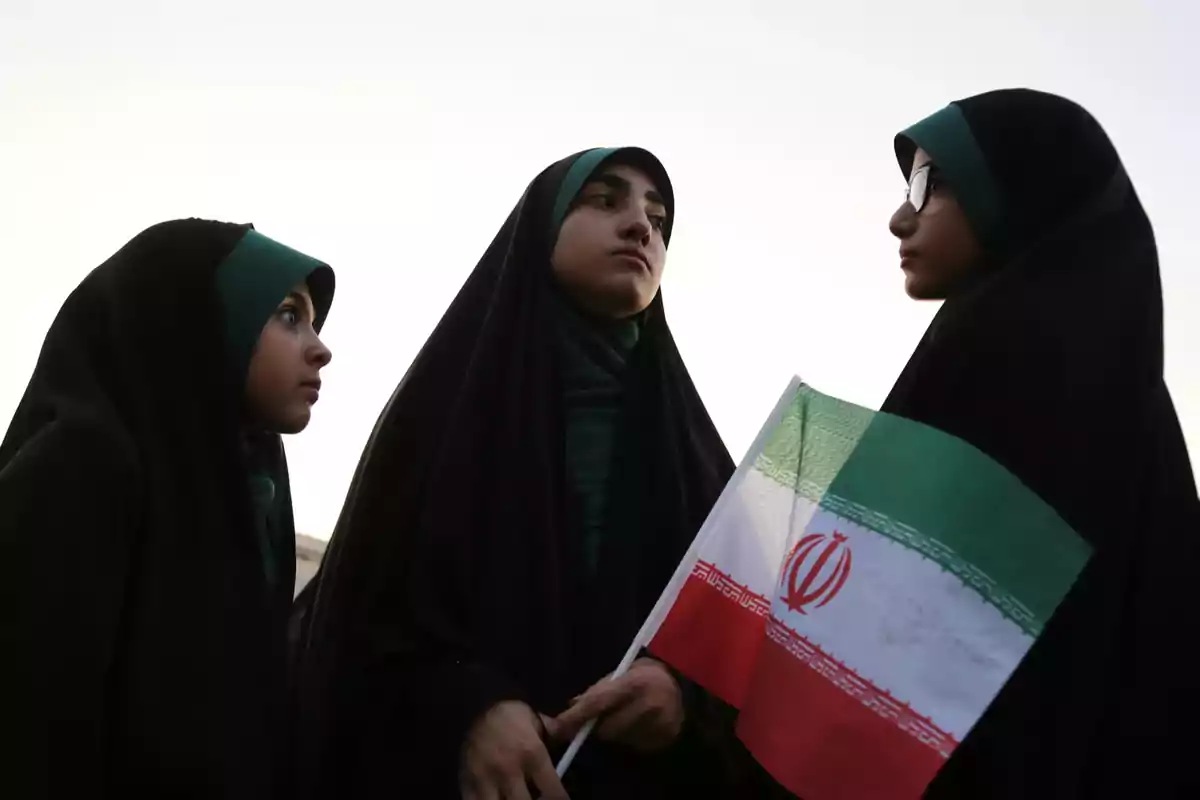 Tres personas con hiyab negro y pañuelos verdes, una de ellas sostiene una bandera de Irán.