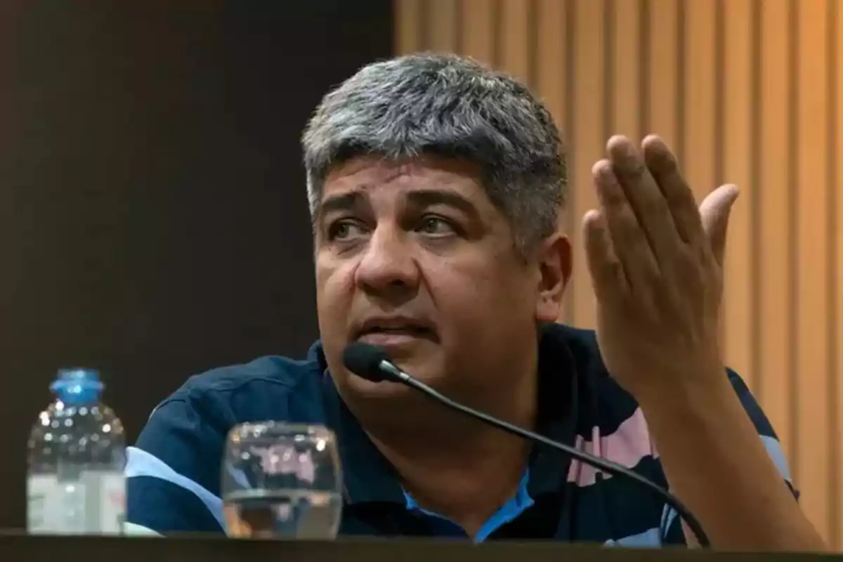 Un hombre con cabello canoso y barba incipiente está hablando en un micrófono mientras gesticula con la mano derecha, frente a él hay una botella de agua y un vaso.