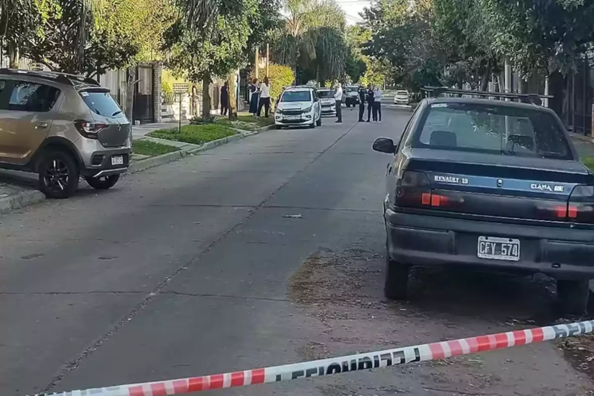 Una calle residencial con varios autos estacionados y una cinta de seguridad en primer plano, mientras algunas personas y un vehículo policial se encuentran al fondo.