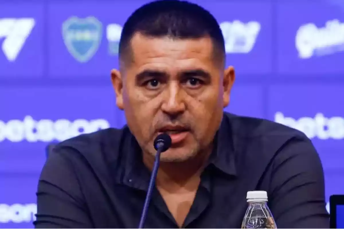 Man speaking at a press conference with a blue background and logos.