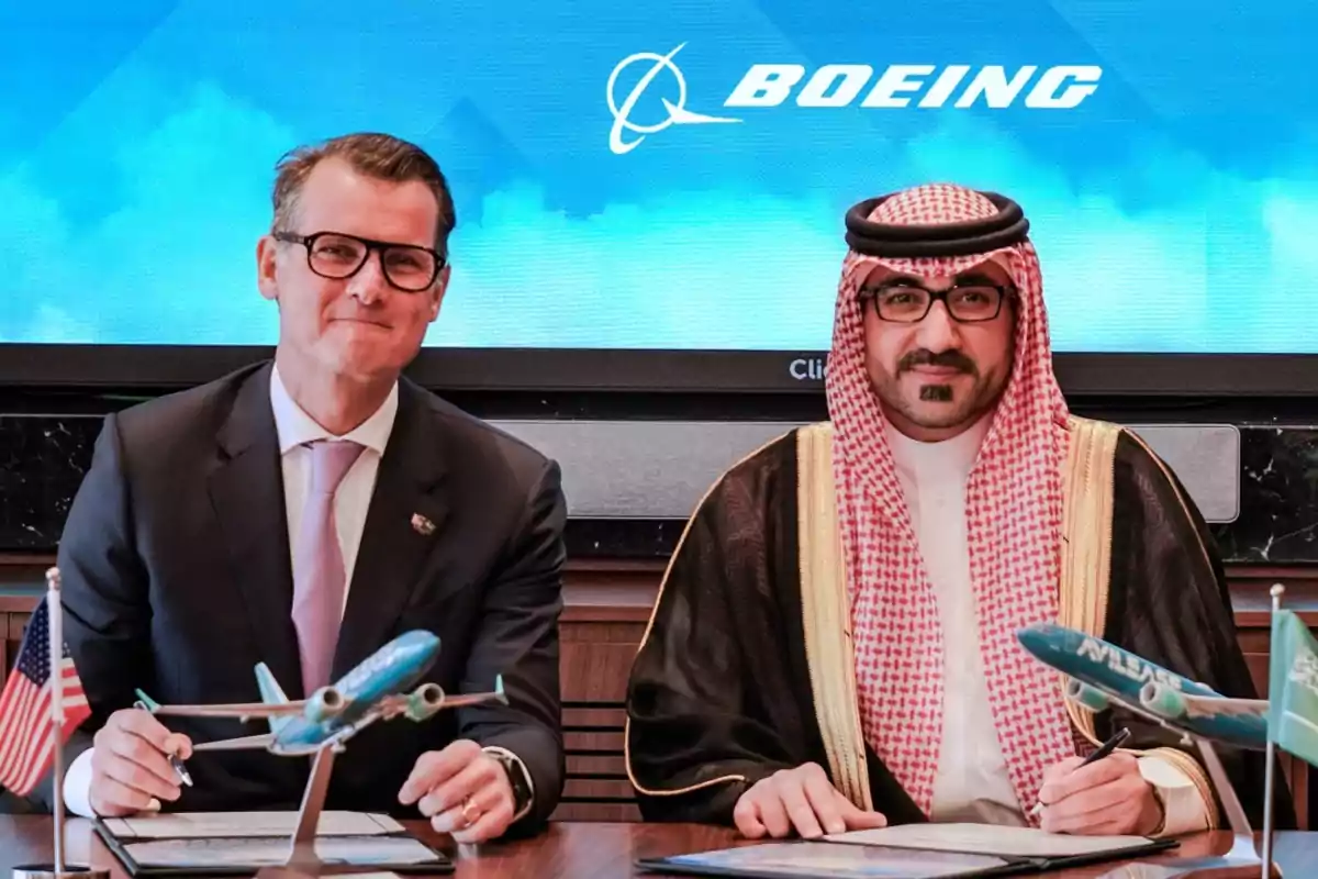 Dos hombres sentados en una mesa con maquetas de aviones y banderas, con un logotipo de Boeing en el fondo. Dos hombres sentados en una mesa con maquetas de aviones y banderas, con un logotipo de Boeing en el fondo.