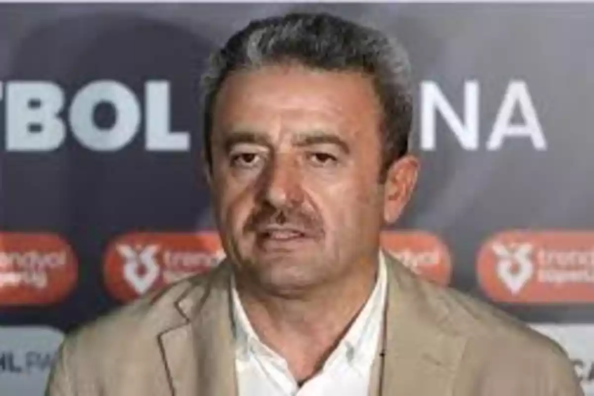 Un hombre con bigote y cabello canoso en una conferencia de prensa con un fondo que tiene texto parcialmente visible. Un hombre con bigote y cabello canoso en una conferencia de prensa con un fondo que tiene texto parcialmente visible.