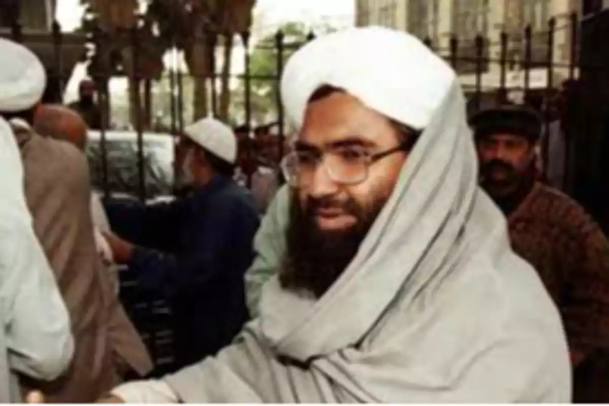 Un hombre con barba y gafas, vestido con una túnica y un turbante blanco, rodeado de otras personas en un entorno urbano. Un hombre con barba y gafas, vestido con una túnica y un turbante blanco, rodeado de otras personas en un entorno urbano.