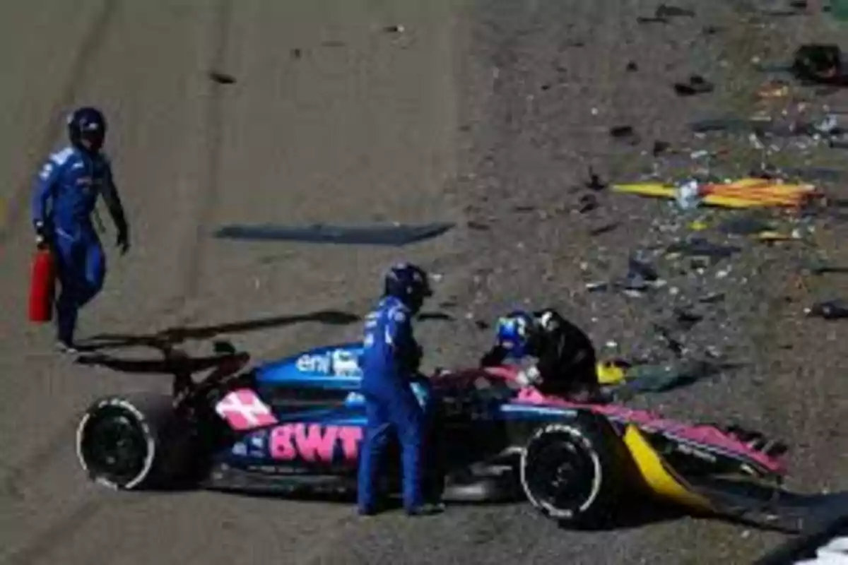 A race car with damage on the track surrounded by people in blue suits. A race car with damage on the track surrounded by people in blue suits.