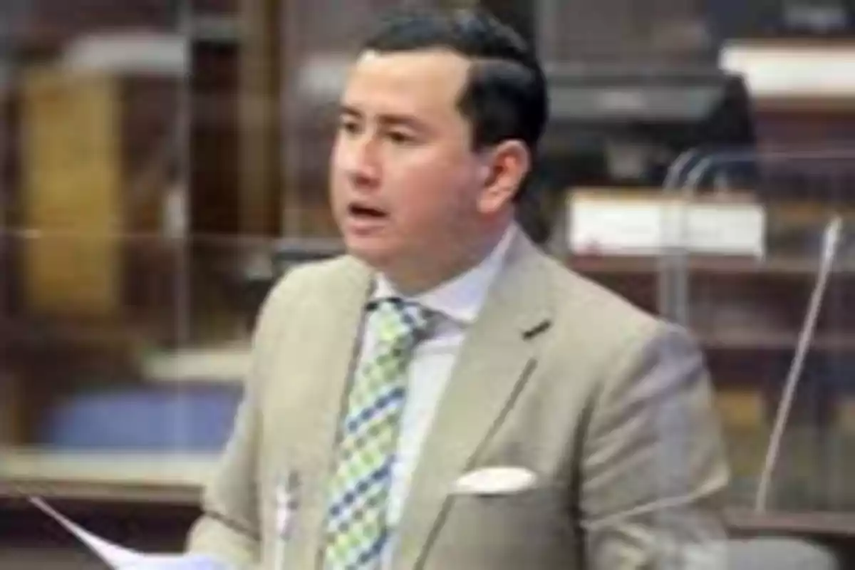 Short dark-haired man wearing a beige jacket and a green plaid tie speaking in a formal setting Short dark-haired man wearing a beige jacket and a green plaid tie speaking in a formal setting