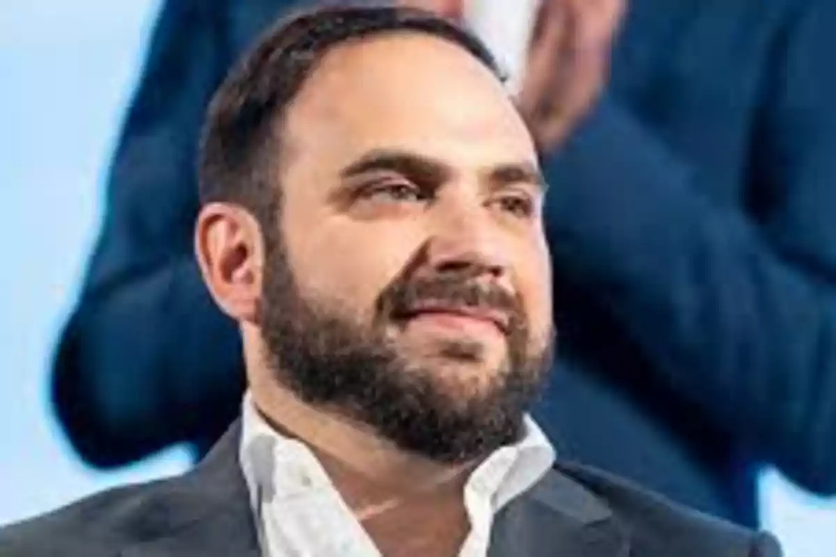 Bearded man with short hair wearing a white jersey and dark jacket in the foreground