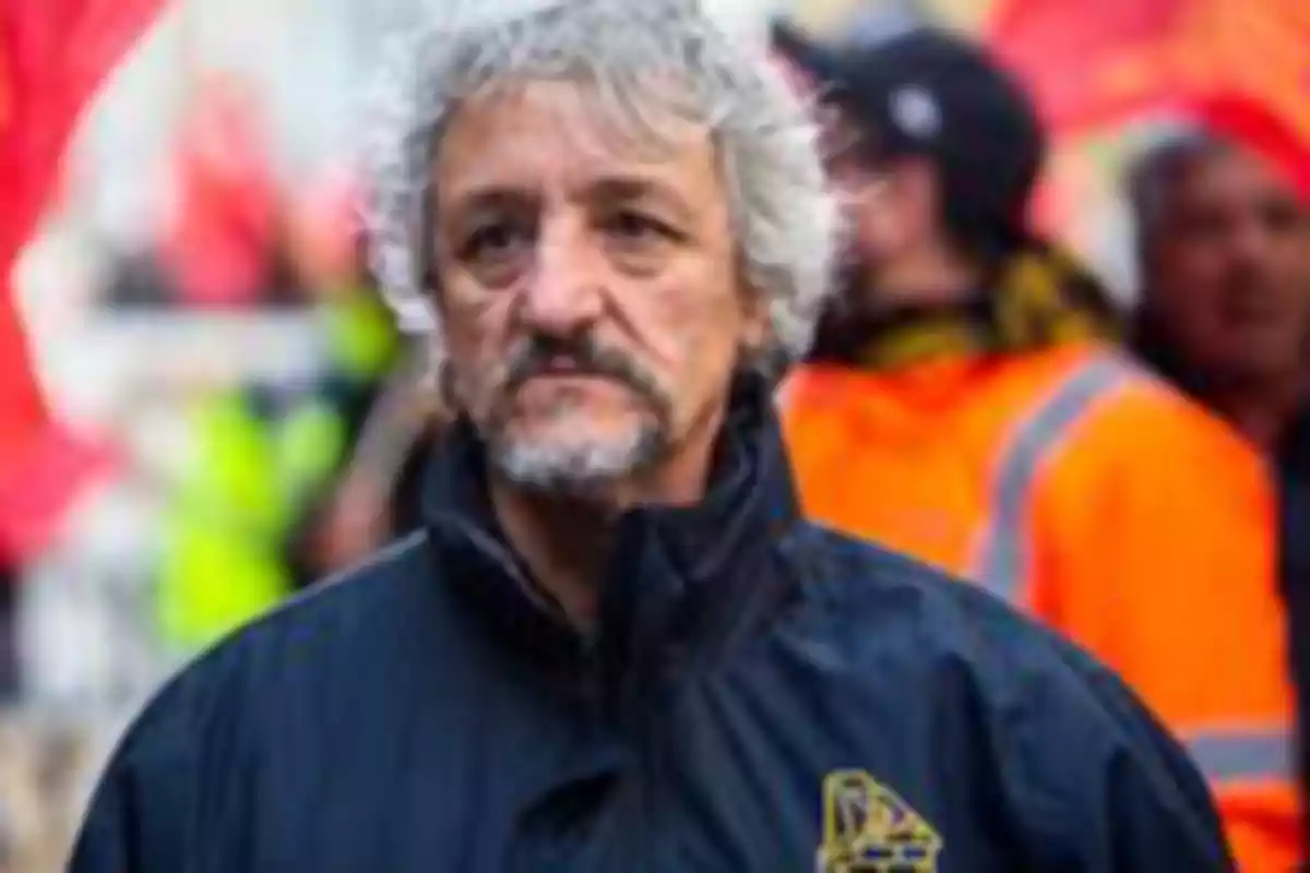Gray-haired, bearded man with a serious expression at a protest, with people and flags blurred in the background Gray-haired, bearded man with a serious expression at a protest, with people and flags blurred in the background