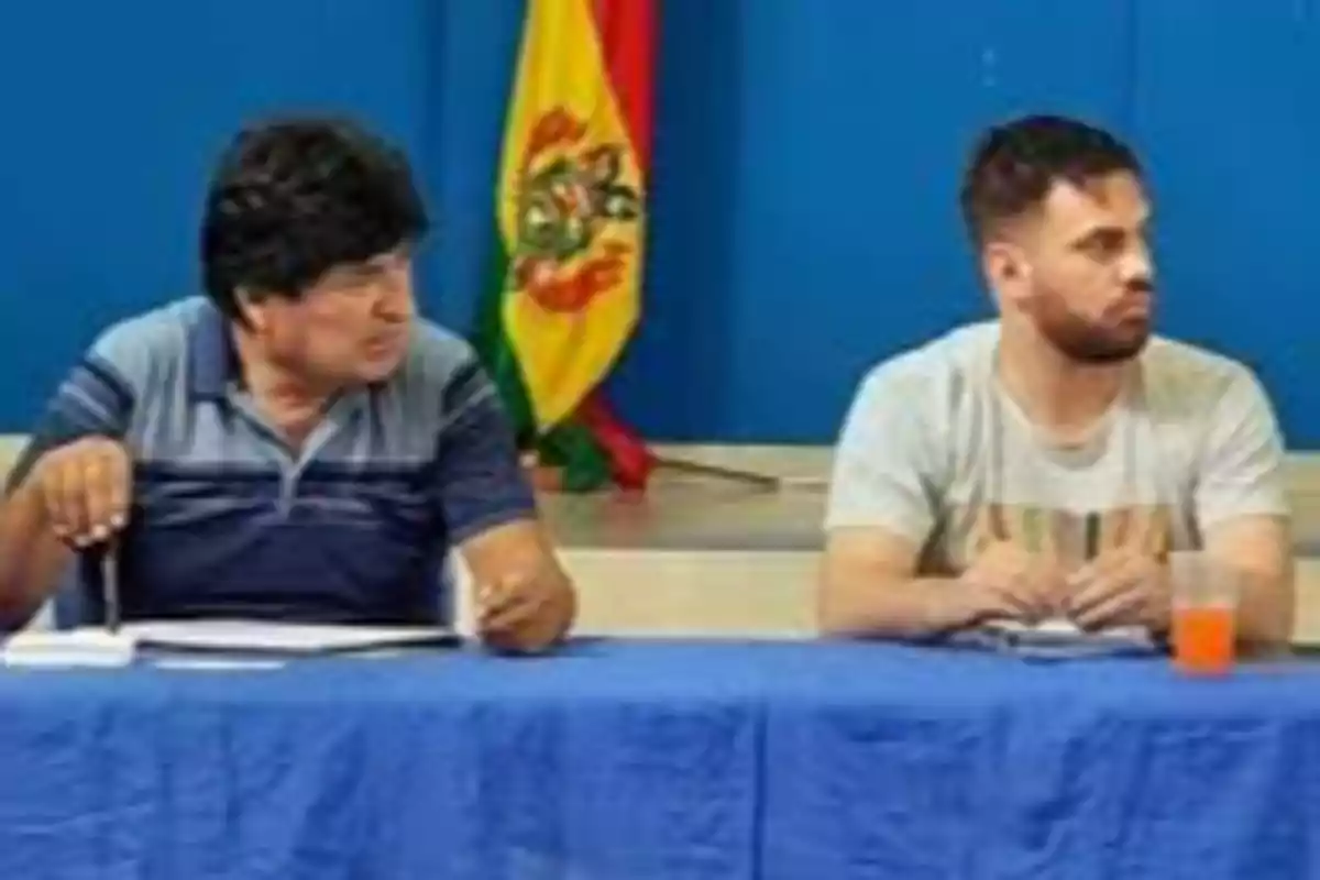 Dos personas sentadas en una mesa cubierta con un mantel azul, al fondo una bandera de Bolivia y una pared azul Dos personas sentadas en una mesa cubierta con un mantel azul, al fondo una bandera de Bolivia y una pared azul