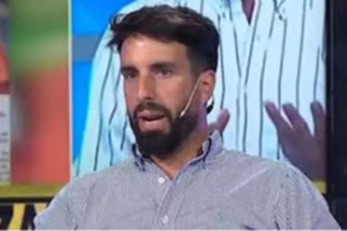 Hombre con barba y camisa a rayas hablando en un programa de televisión con un micrófono en la oreja.