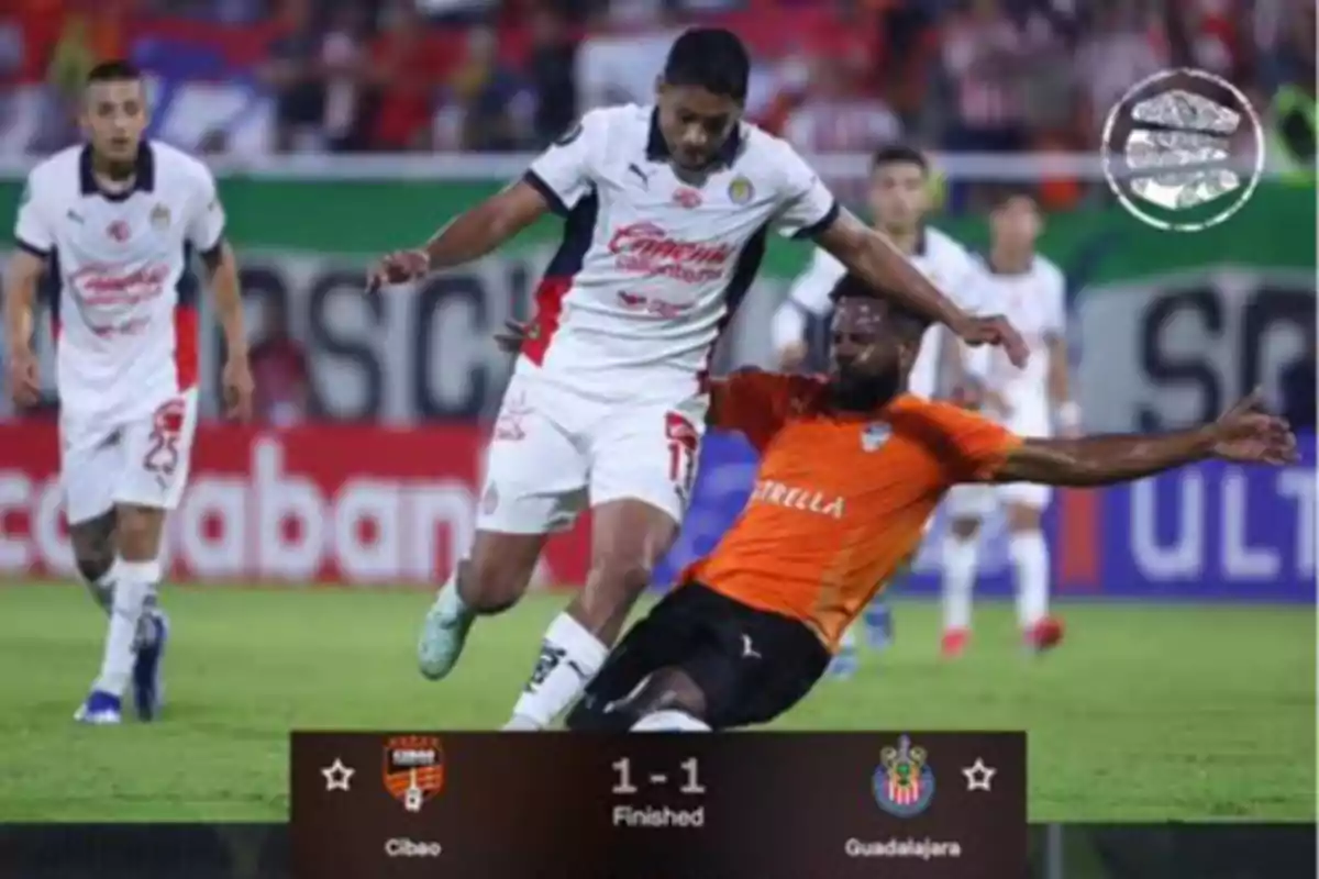 Un jugador de fútbol con uniforme blanco intenta avanzar mientras otro jugador con uniforme naranja se desliza para quitarle el balón en un partido que terminó 1-1 entre Cibao y Guadalajara.