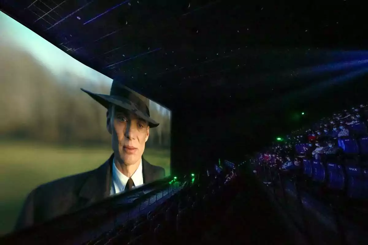 A dark movie theater with a large screen showing a man in a hat and suit, while the audience watches from their seats. A dark movie theater with a large screen showing a man in a hat and suit, while the audience watches from their seats.