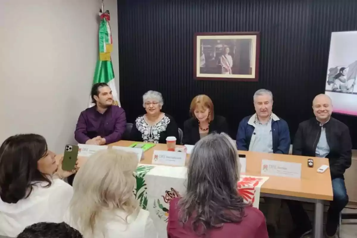 Un grupo de personas está sentado alrededor de una mesa en una reunión, con una bandera de México en la pared y un cuadro en el fondo.