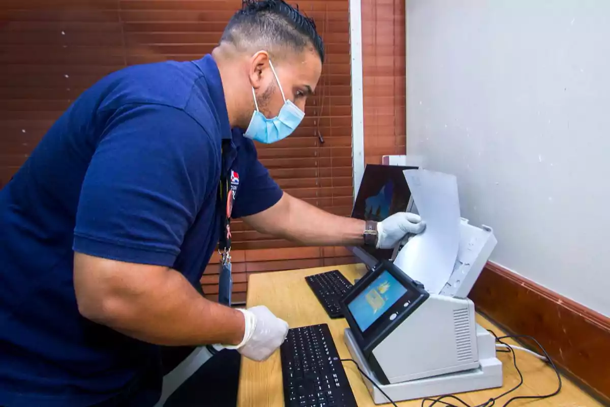 Hombre con mascarilla y guantes digitalizando documentos en una oficina usando un escáner y una computadora Hombre con mascarilla y guantes digitalizando documentos en una oficina usando un escáner y una computadora