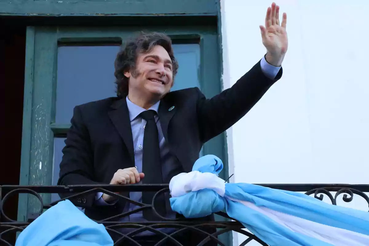 Un hombre sonriente con traje oscuro y corbata saluda desde un balcón decorado con una bandera celeste y blanca.