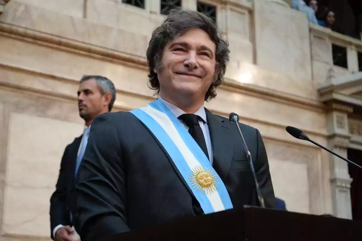 Un hombre con traje y banda presidencial sonríe mientras está de pie frente a un micrófono en un edificio gubernamental.