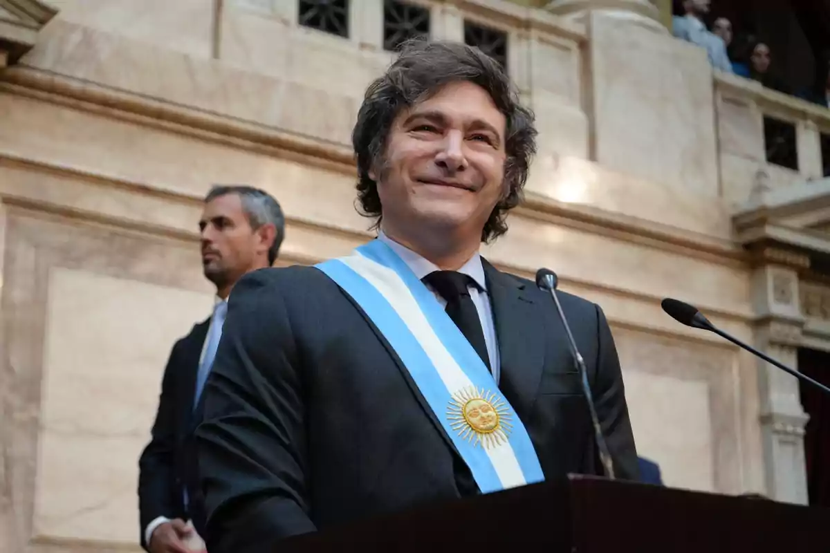 Un hombre con traje y banda presidencial sonríe mientras está de pie frente a un micrófono en un edificio con arquitectura clásica.