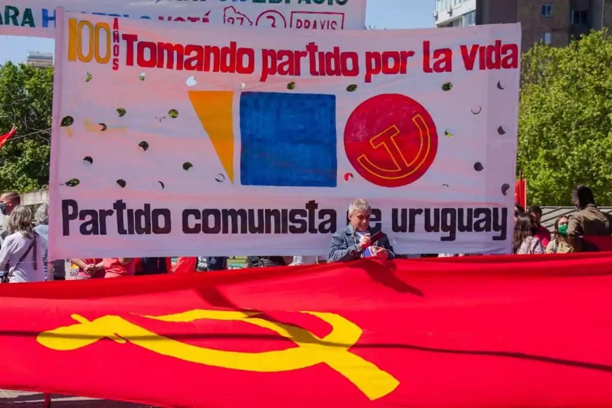 A group of people holds a banner of the Communist Party of Uruguay with the slogan "Taking a stand for life" and a communist symbol at an outdoor event.