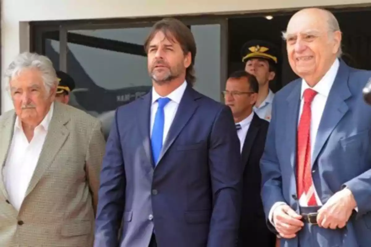 Tres hombres de pie con trajes formales en un evento al aire libre.