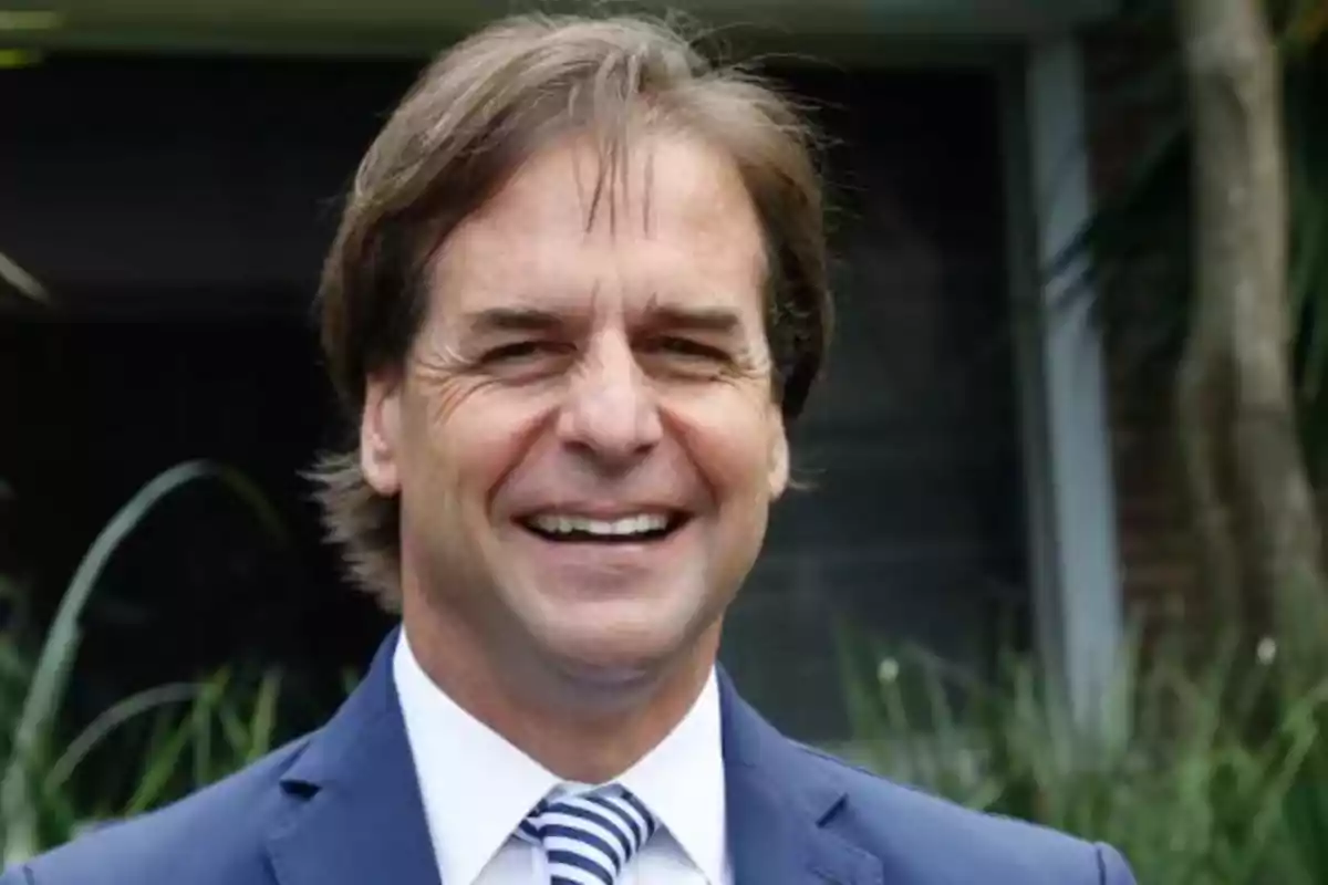 Hombre sonriendo con traje azul y corbata a rayas en un entorno al aire libre.