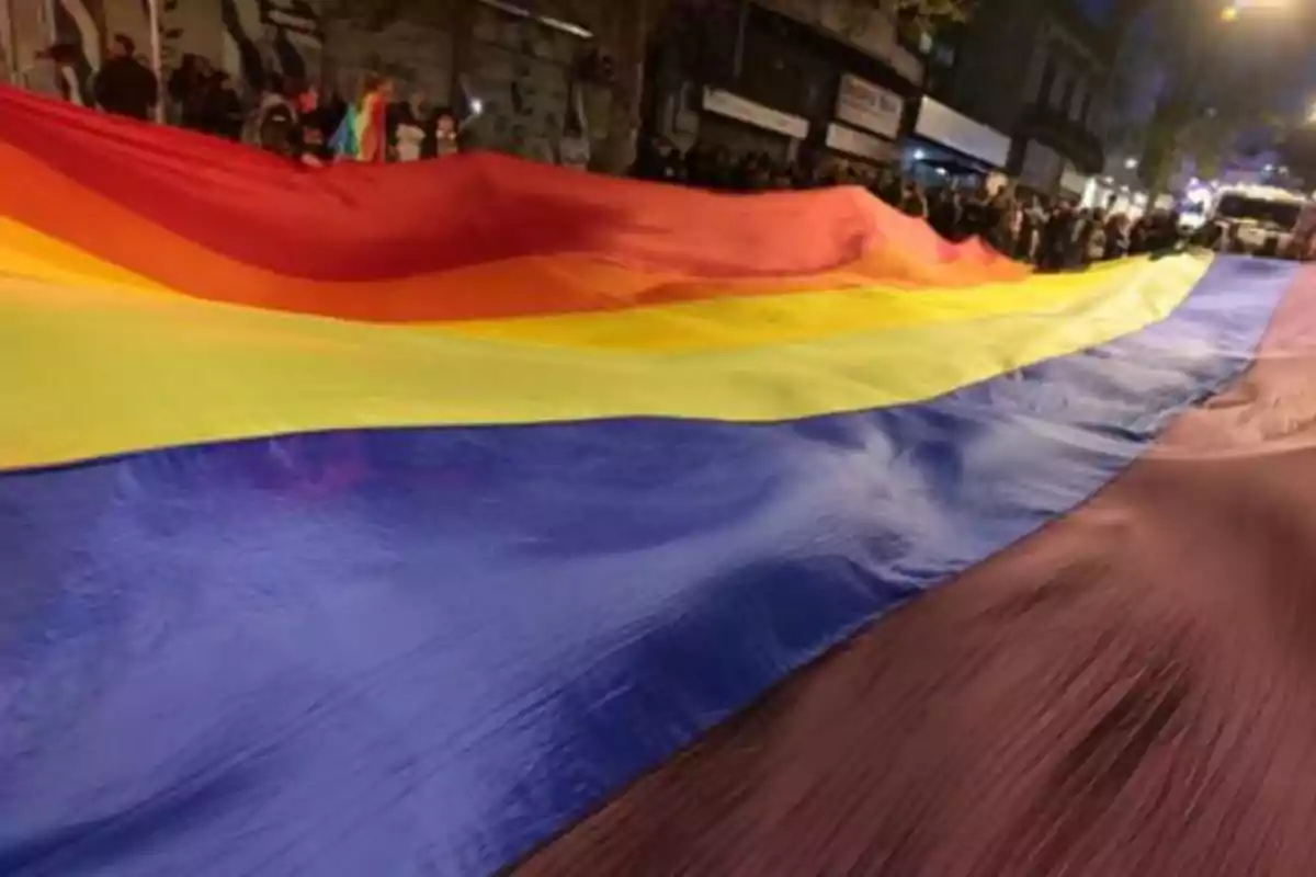 Una gran bandera con los colores del arcoíris es sostenida por personas en una calle durante la noche.