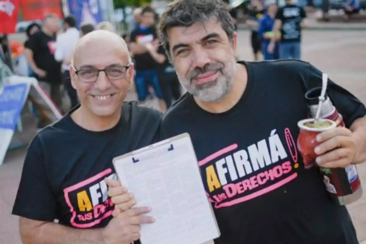 Dos personas sonríen a la cámara mientras una sostiene un mate y la otra un portapapeles, ambas llevan camisetas con un mensaje sobre firmar por los derechos.