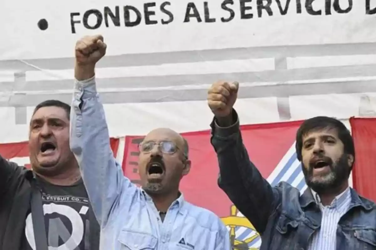 Tres personas levantan el puño en señal de protesta frente a una pancarta.