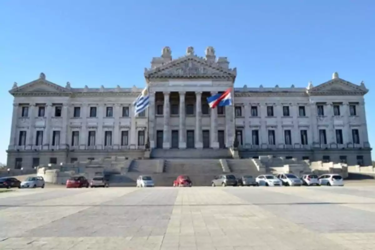 Edificio del Palacio Legislativo de Uruguay con banderas ondeando al frente y varios coches estacionados en la explanada.