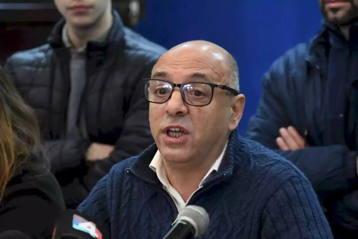 Un hombre con gafas y suéter azul habla frente a un micrófono en una conferencia de prensa, rodeado de otras personas.