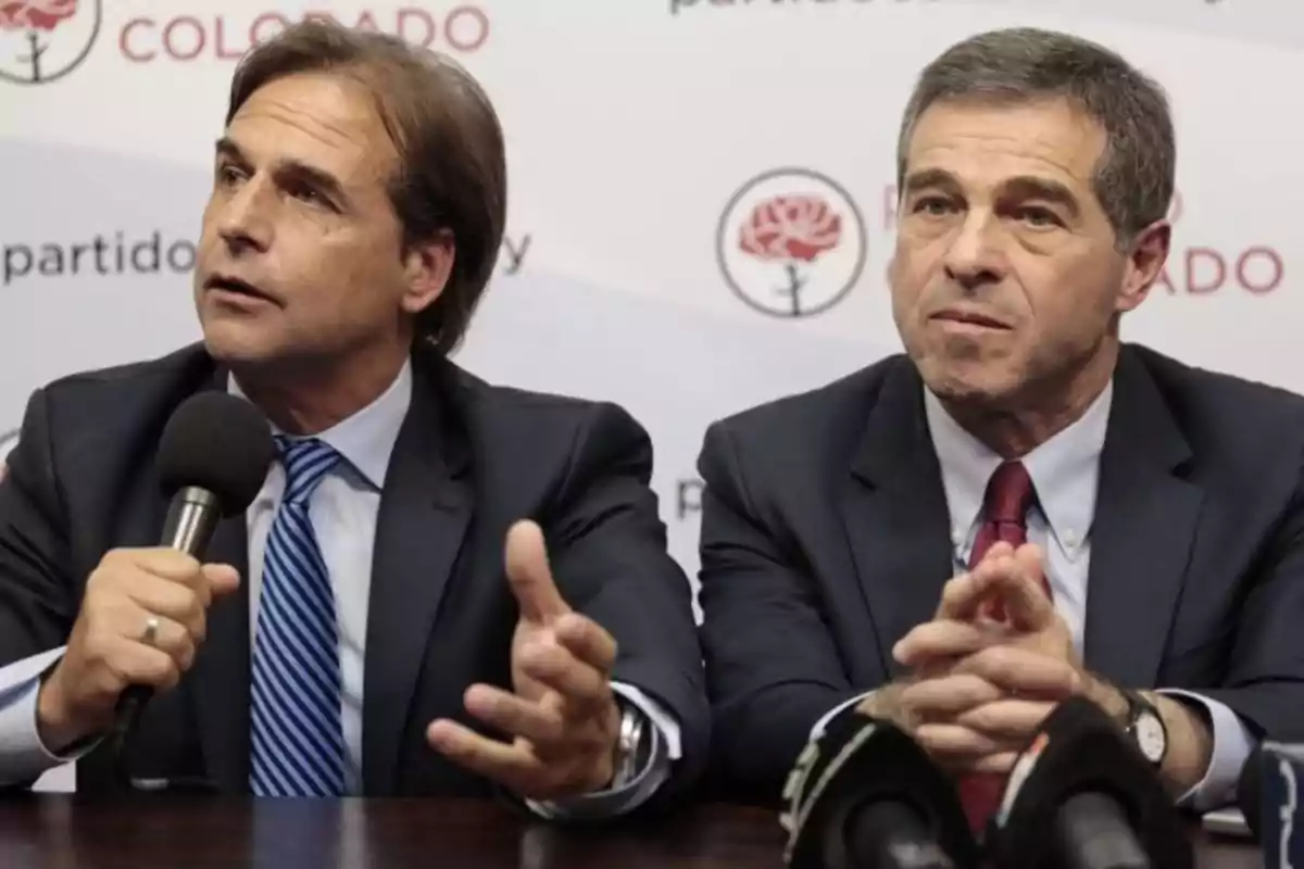 Dos hombres en traje hablando en una conferencia de prensa con micrófonos frente a ellos y un fondo que menciona "partido colorado".