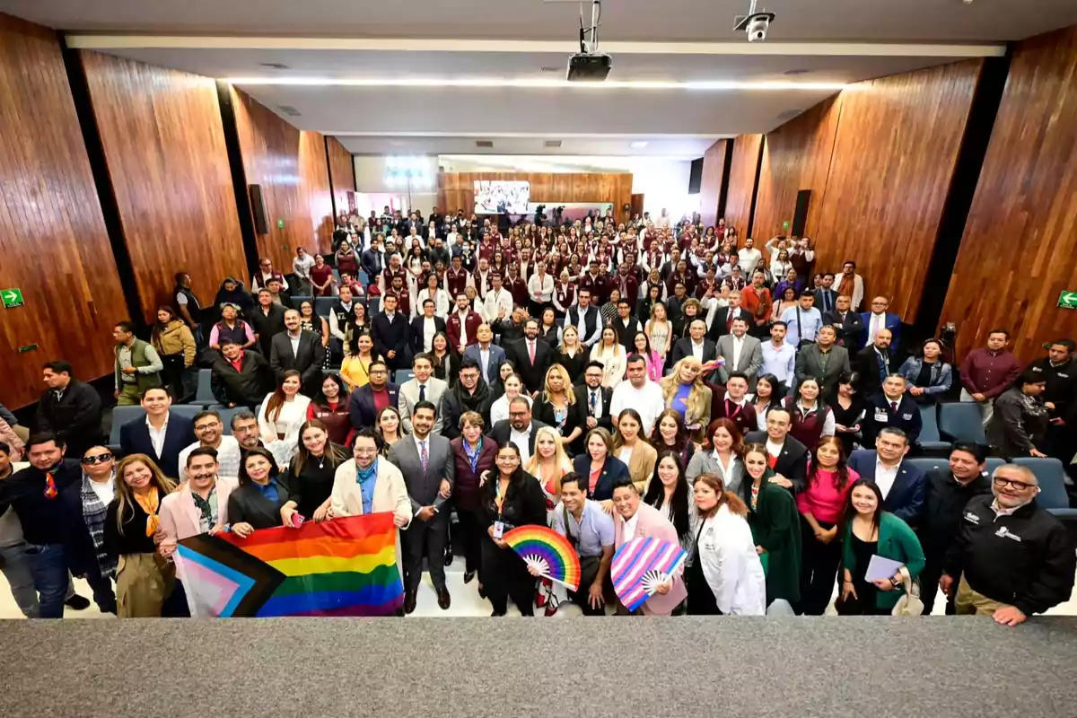 Un grupo numeroso de personas posando en un auditorio, algunas sosteniendo banderas y abanicos con los colores del arcoíris.