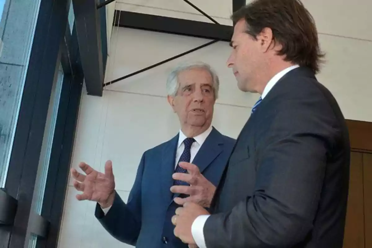 Dos hombres de traje conversan junto a una ventana en un entorno interior.