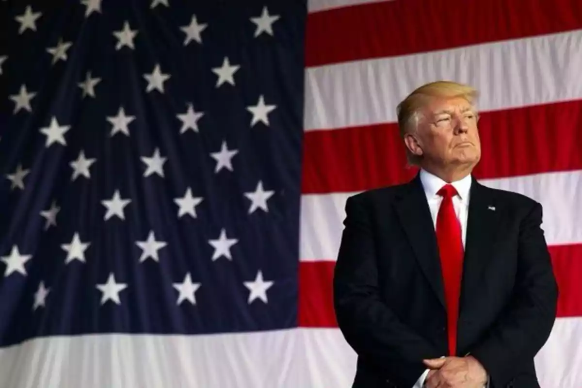 Un hombre de pie con traje oscuro y corbata roja frente a una gran bandera de Estados Unidos.