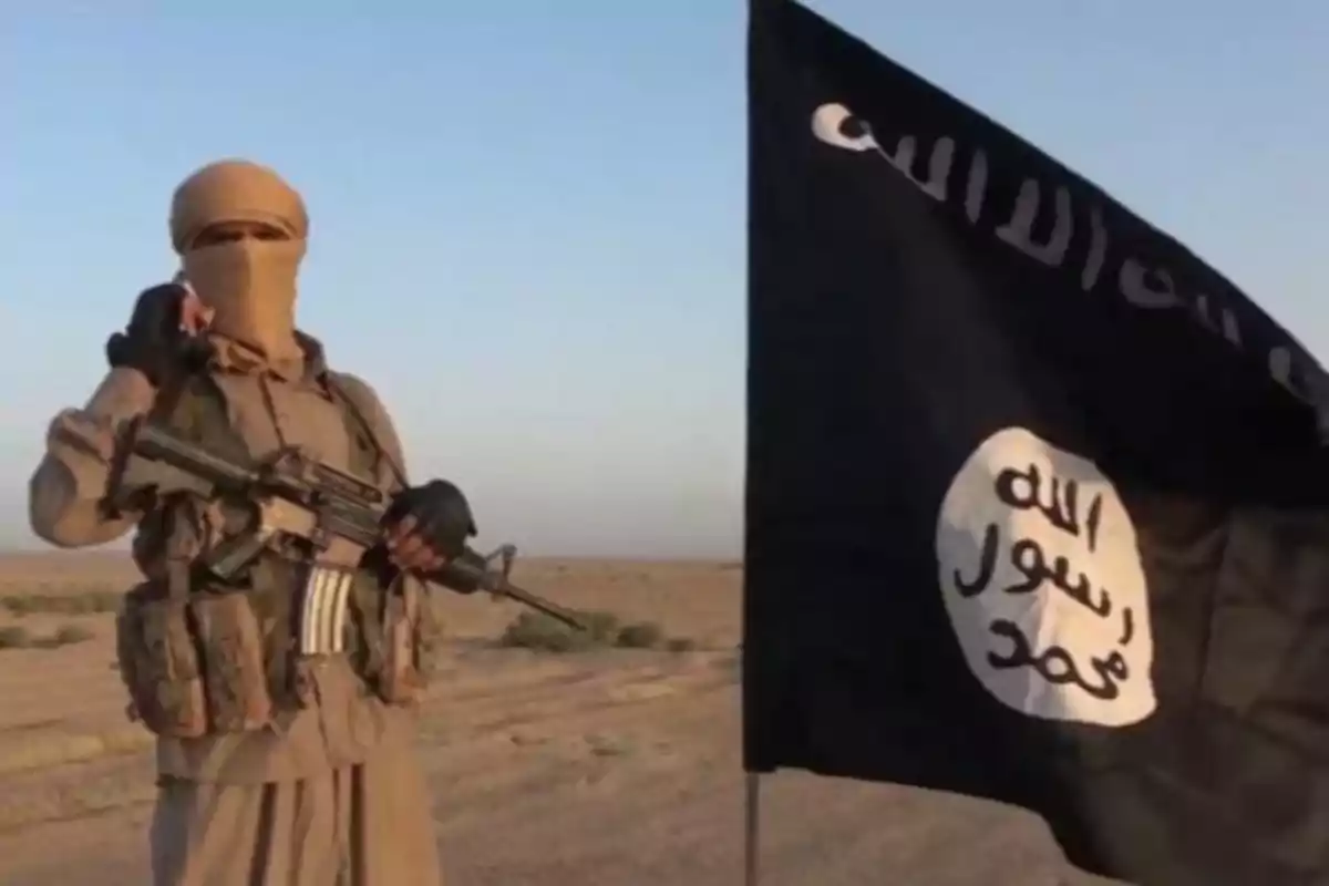 Una persona armada con el rostro cubierto junto a una bandera negra en un paisaje desértico.