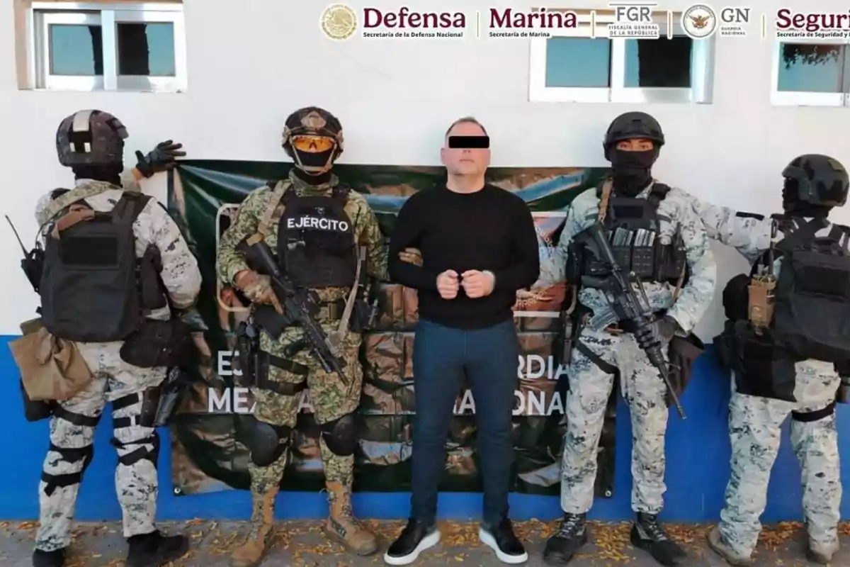 A group of armed military personnel escorts a man with covered eyes in front of an institutional sign.