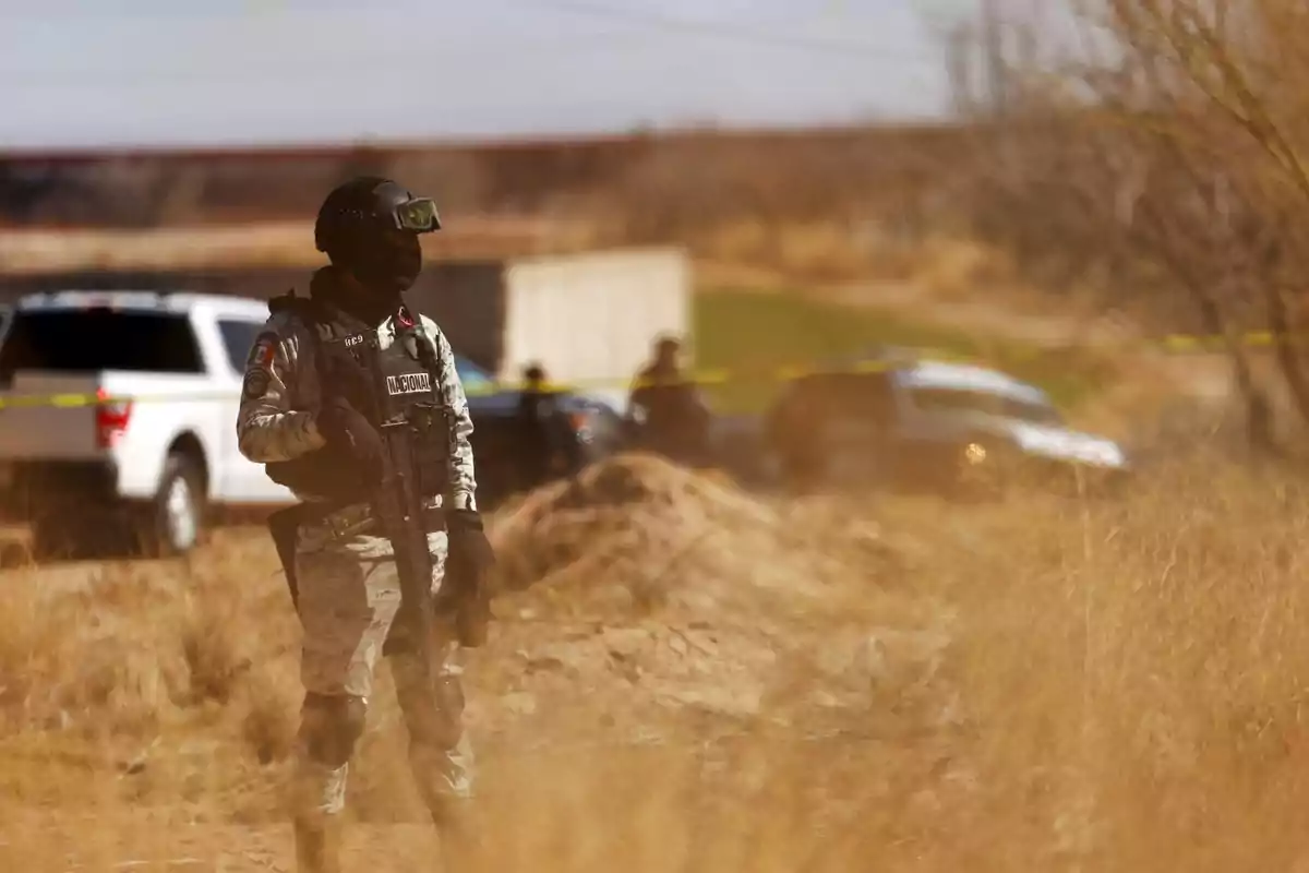 Un miembro de la Guardia Nacional de pie en un área rural con vehículos y cinta de seguridad al fondo.