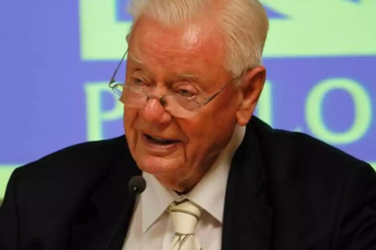 An older man with white hair and glasses speaking in front of a microphone.