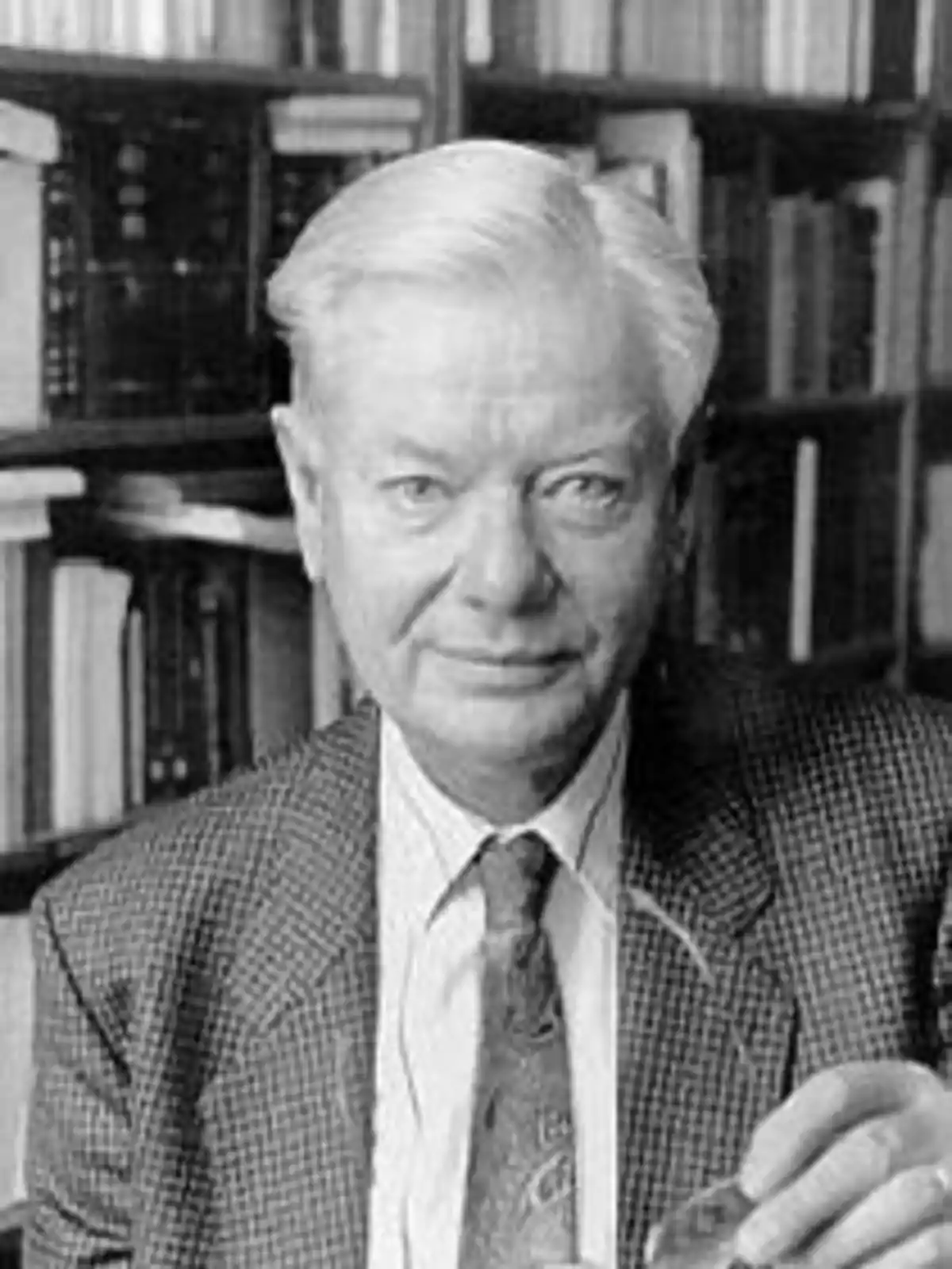 An older man with gray hair and a suit, sitting in front of a bookshelf full of books.