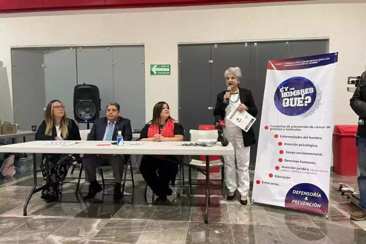 Un grupo de personas está sentado en una mesa durante un evento, mientras una mujer de pie sostiene un micrófono y un folleto, junto a un cartel que promueve la atención a la salud masculina y la prevención de enfermedades.