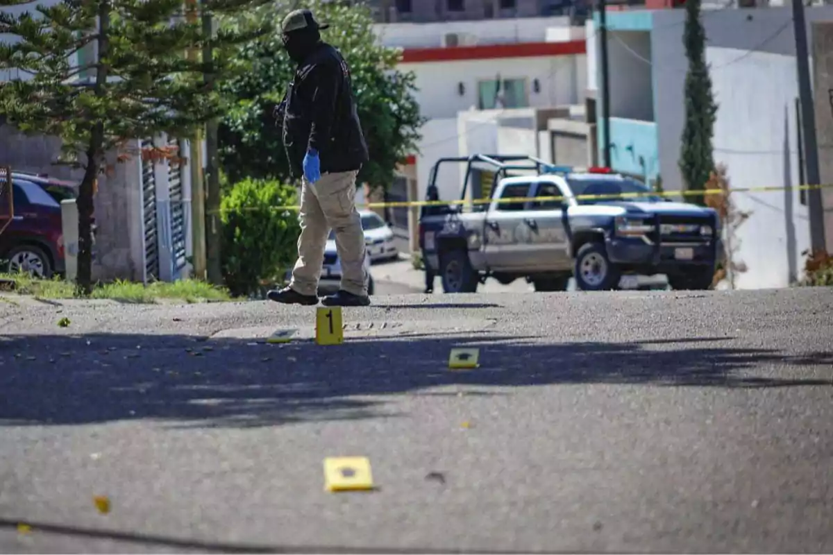 Una persona con guantes y mascarilla camina por una calle con marcadores de evidencia numerados, mientras una camioneta de policía está estacionada al fondo.