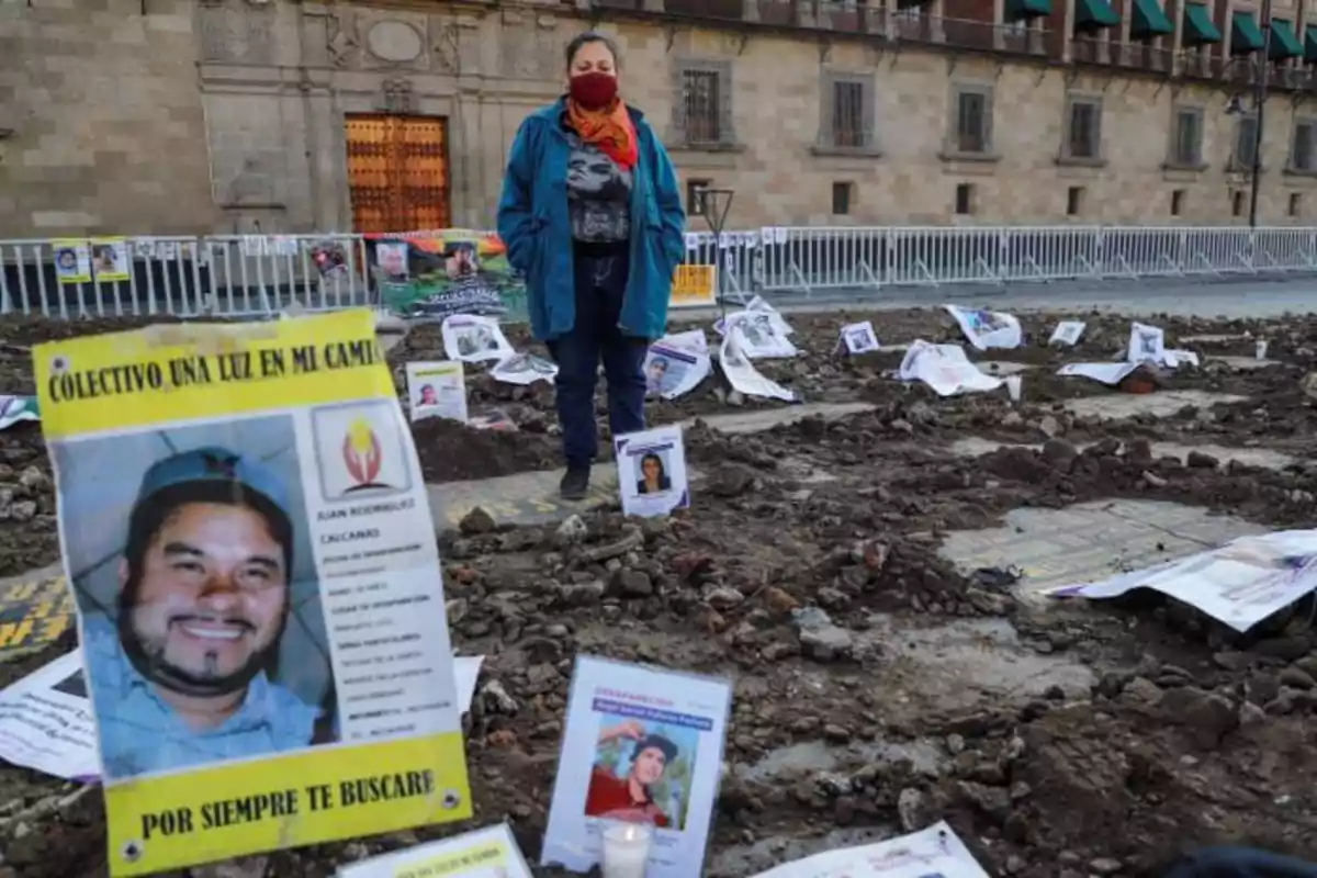 Una persona con mascarilla está de pie en un área con tierra removida, rodeada de carteles y fotografías de personas desaparecidas, frente a un edificio histórico.