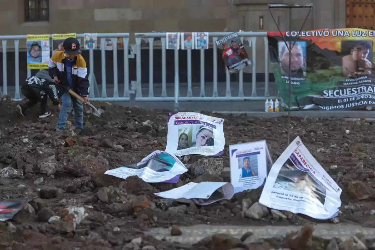 Un niño con gorra y chaqueta cava en un terreno lleno de tierra y piedras, mientras que en el suelo hay carteles con fotos de personas desaparecidas y al fondo se ven pancartas colgadas en una valla.