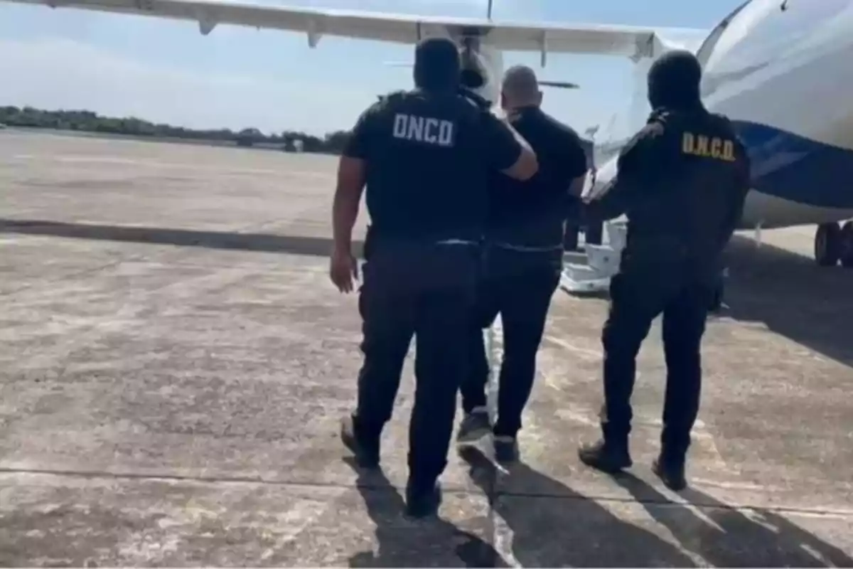 Un grupo de agentes escolta a una persona hacia un avión en una pista de aterrizaje.